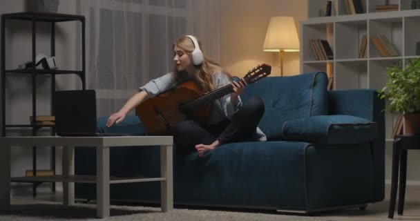 Giovane donna sta imparando a suonare la chitarra online, visualizzazione tutorial sul computer portatile, hobby nel tempo libero alla sera — Video Stock