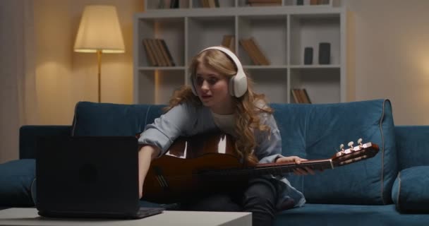 젊은 여성은 인터넷으로 기타를 연주하기 위해 공부하고 있습니다. 노트북 화면으로 비디오 튜토리얼을 켜고, 저녁에 집에 앉아서 — 비디오