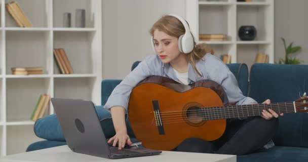 Mulher bonita com fones de ouvido está ligando vídeo lição como jogar guitarra na internet no notebook, segurando guitarra e arrancando cordas — Vídeo de Stock
