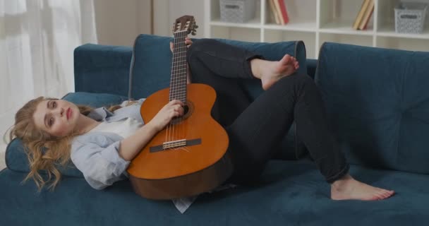 Ontspannen jonge vrouw ligt op de bank in de woonkamer en spelen gitaar, zorgeloos en romantisch humeur, geïnspireerd beginner muzikant — Stockvideo