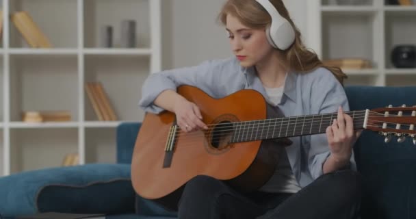 Молодая женщина кантри музыкант играет на акустической гитаре в гостиной своего дома, сидя на диване перед ноутбуком — стоковое видео