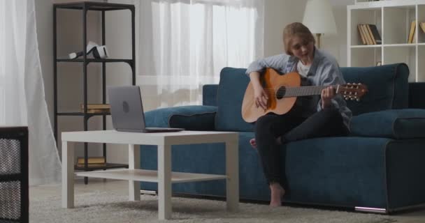 Fiatal női zenész tanul új összhangban gitár lakásban, ül a kanapén hangszeren, játszik húrok — Stock videók