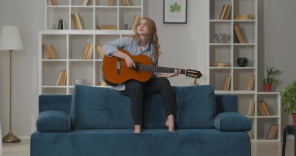 Begåvad ung sångerska kvinna sitter på baksidan av soffan i mysiga vardagsrum och spela sång på gitarr, kul och musik — Stockvideo