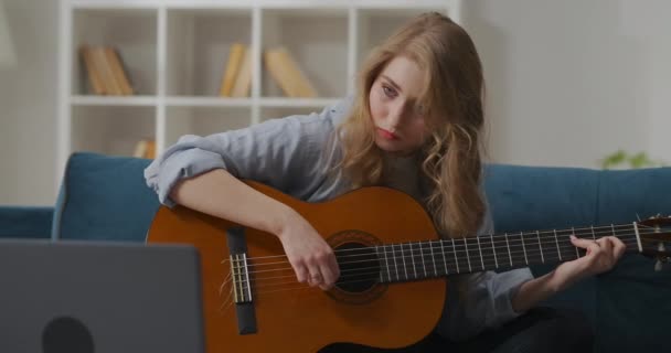 Bastante joven mujer recogiendo la melodía en la guitarra, sentado solo en la sala de estar, pasar tiempo en casa por la noche — Vídeo de stock