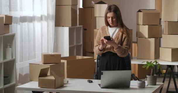 Молодая женщина в своей рабочей квартире с коробками, используя мобильный телефон для общения с курьером — стоковое видео