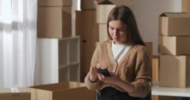 Dairesinde karton kutular olan bir depo. Kadın akıllı telefon kullanıyor. Müşterilerle teslimat hakkında iletişim kuruyor.