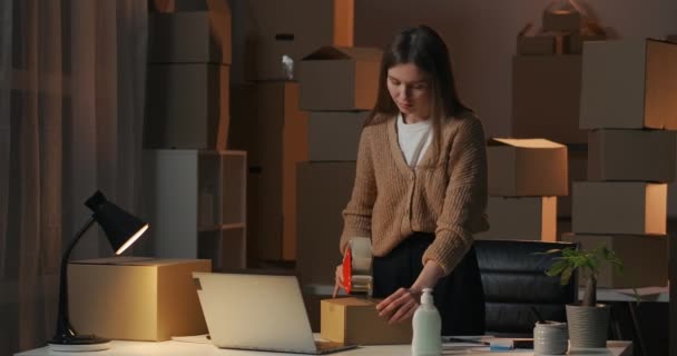 Giovane imprenditore donna sta sigillando scatola con merci per la consegna da roll-on tape dispenser di notte nel suo appartamento, ritratto medio — Video Stock