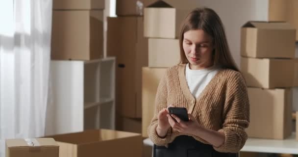 Jonge vrouw is eigenaar van kleine bedrijven, staan in magazijn in appartement en het bekijken van foto 's van goederen in smartphone — Stockvideo