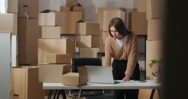 Young woman is chatting by video call in laptop looking on screen and web camera in her home warehouse with boxes — Stock Video