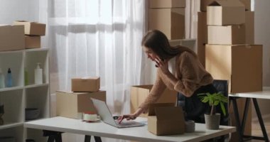 Meşgul kadın müşterileriyle cep telefonuyla konuşuyor ve bilgisayardaki bilgileri kontrol ediyor, ev deposunda çalışıyor.