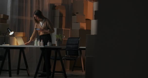 젊은 사업가의 아파트에 화장품 상자가 있는 집 창고에서, 한 부인이 저녁에 혼자 일하고 있다 — 비디오
