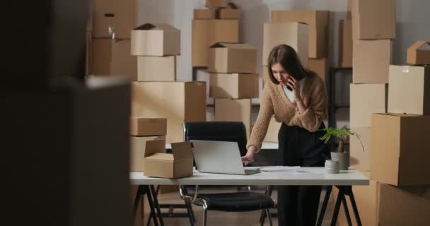 Customer service of small delivery company, woman is consulting clients by phone and filling in order in laptop, portrait in home warehouse — Stock Video
