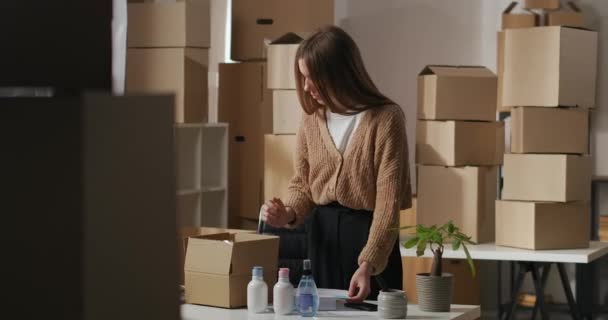 Взрослая женщина распаковывает коробку с косметикой на маленьком складе в квартире, проверяет порядок в документе, малый бизнес и предпринимательство — стоковое видео