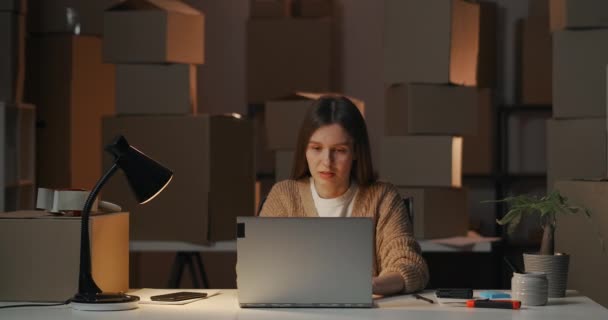 Retrato de una joven empresaria que trabaja en su almacén doméstico con cajas, sentada a la mesa con portátil por la noche, terminando el trabajo — Vídeos de Stock