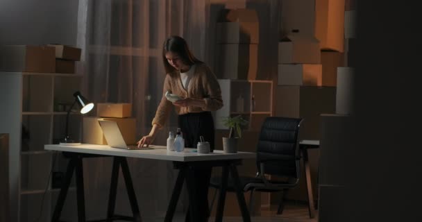 Επιχειρηματίας εργάζεται τη νύχτα στην αποθήκη του σπιτιού της με κουτιά, διαβάζοντας πληροφορίες για τα νέα καλλυντικά προϊόντα — Αρχείο Βίντεο