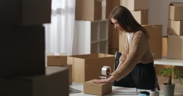 Vrouwelijke werknemer van kleine binnenlandse magazijn maakt gebruik van roll-on tape dispenser voor verpakking doos met goederen, verpakkingsproducten — Stockvideo