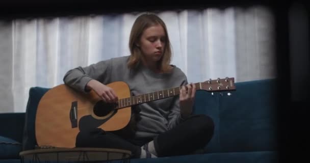 Een tiener in een grijs sweatshirt speelt een lyrische melodie op de gitaar. Zit op een blauwe bank in een comfortabele lotuspositie. De zon schijnt vanuit het raam. Schieten in beweging. — Stockvideo