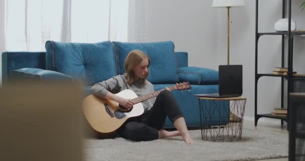 La chica se sienta en el suelo de la sala de estar con la espalda contra el borde del sofá. El músico domina las habilidades de tocar la guitarra. — Vídeo de stock