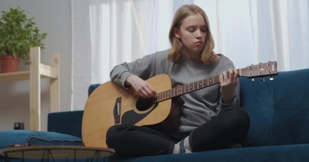 Un adolescente con una sudadera gris toca una melodía lírica en la guitarra. Se sienta en un sofá azul en una cómoda posición de loto. El sol brilla desde la ventana. Tiro medio. — Vídeo de stock