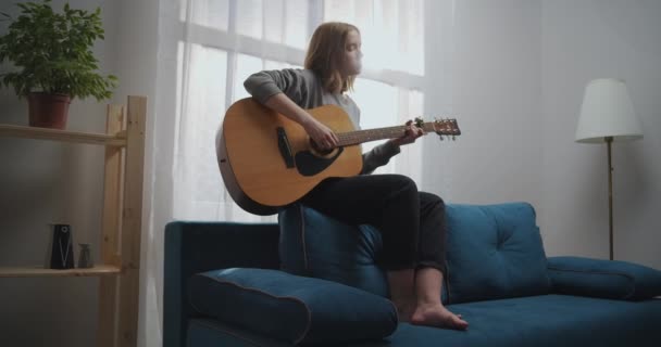 La chica toca la guitarra en la parte posterior del sofá azul. La mujer crea música en el interior. El artista toca la guitarra acústica. El músico compone una melodía. Un adolescente entrena en un instrumento musical. — Vídeos de Stock