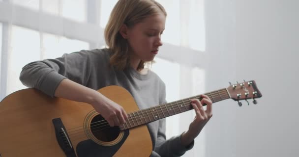 Un adolescente toca la guitarra en una sala de estar blanca con hermosa luz del sol. La mujer compone música en el interior. Una chica está practicando un instrumento musical sola durante una pandemia. — Vídeo de stock