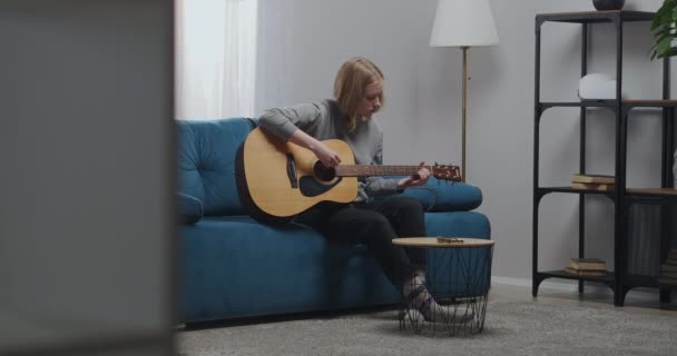 Una joven toca una guitarra acústica mientras está sentada en un sofá azul suave en la sala de estar de su casa durante el día. — Vídeos de Stock