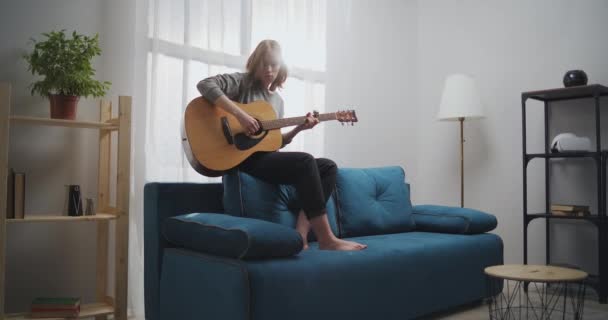Het meisje speelt gitaar op de bank. Een vrouw componeert muziek in een lichte woonkamer met prachtig licht. De artiest speelt de klassieke gitaar. De muzikant componeert een nieuwe melodie. — Stockvideo