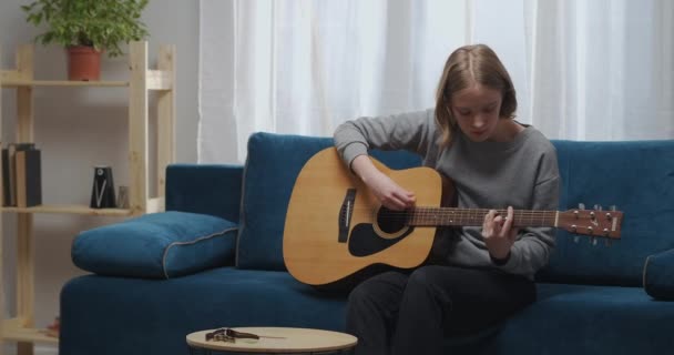 En ung flicka lär sig spela gitarr genom att slå till medan hon sitter på soffan i sitt rum. Medelhög skott. — Stockvideo