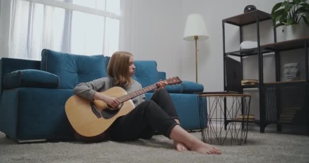 Gyönyörű nő néz online leckét, miközben ül a padlón a szobában. A lány elszigetelten tanul gitározni. Egy törekvő zenész megtanul hangszeren játszani laptoppal. — Stock videók