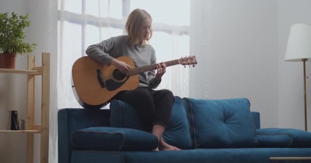 Una giovane ragazza con una felpa grigia e pantaloni neri compone nuova musica su una chitarra acustica. Si siede sul retro di un morbido divano a piedi nudi in un luminoso soggiorno. — Video Stock