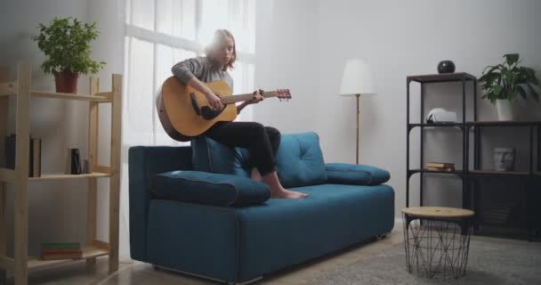 The girl plays the guitar on the couch. A woman alone creates music in a white living room. The artist plays the classical guitar. The musician composes a dreary melody. Long short. — Stok Video