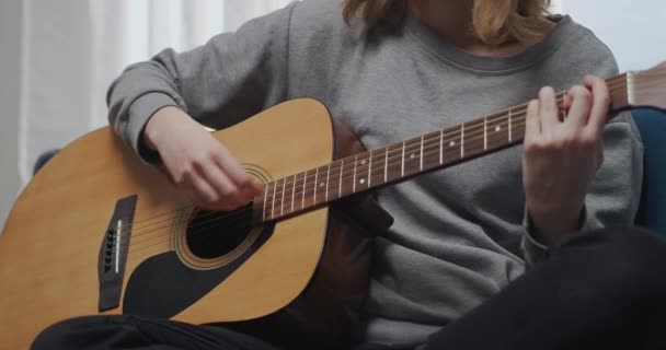 Ein Teenager im grauen Sweatshirt spielt eine lyrische Melodie auf der Gitarre. Sitzt auf einem blauen Sofa in einer bequemen Lotusposition. Die Sonne scheint aus dem Fenster. Nahaufnahme — Stockvideo