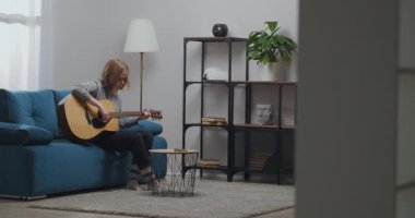 Genç kız gitarda güzel bir melodi çalar. Sarı saçı kısa saç kesimi. Odanın içi İskandinav tarzında yapılmış..