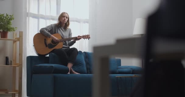 En tonårstjej i grå tröja och svarta byxor spelar sin favoritmusik på en akustisk gitarr. Sitter på baksidan av en blå soffa med bara fötter i ett modernt vardagsrum. Fotografering i rörelse. — Stockvideo