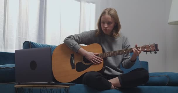 Een jong meisje zit op de bank in de lotuspositie in de woonkamer. Een vrouw leert alleen gitaar spelen. Ze kijkt naar haar eerste online les op haar laptop. — Stockvideo