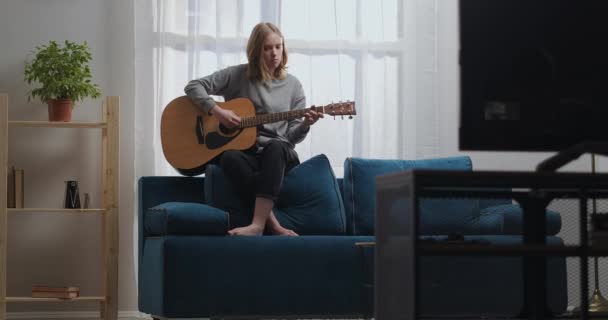 Une adolescente en sweat-shirt gris et pantalon noir sans chaussettes joue une mélodie calme sur une guitare acoustique. Assis sur le dos d'un canapé bleu. Le soleil brille par la fenêtre derrière. — Video