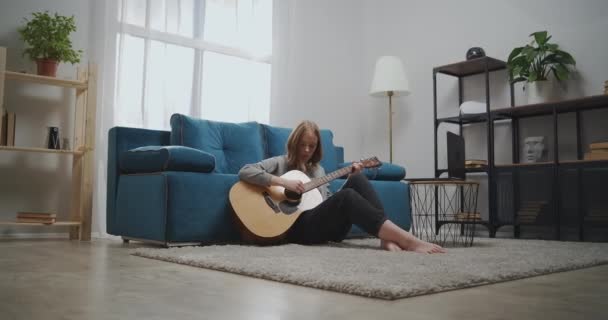 Mooie vrouw kijken online les terwijl zitten op de vloer in de kamer. Meisje leert gitaar spelen. Een beginnende muzikant leert met een laptop een muziekinstrument bespelen. Grote kans. — Stockvideo