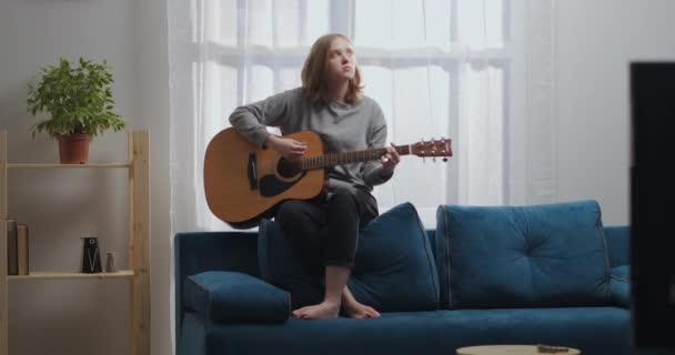 Una adolescente con una sudadera gris y pantalones negros toca una melodía lírica en la guitarra. Se sienta en la parte posterior de un sofá azul. El sol brilla desde la ventana de atrás. — Vídeos de Stock