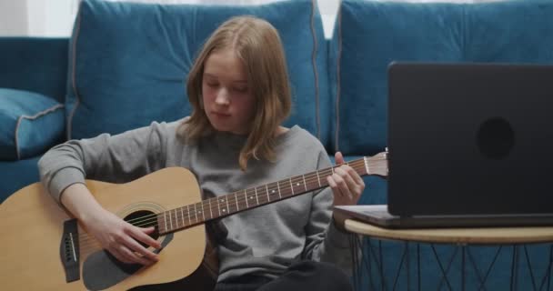 Nastolatek siedzi na podłodze plecami do krawędzi miękkiej sofy w salonie. Muzyk uczy się nowych akordów na gitarze. Strzał z bliska. — Wideo stockowe