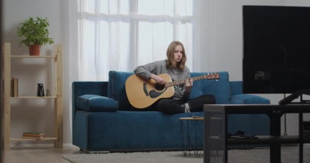 Tini lány otthoni ruhában játszik egy vidám dallam egy új klasszikus gitár ül kényelmes helyzetben egy kék kanapén egy meleg nappaliban — Stock videók