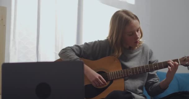Een mooi meisje kijkt naar een online les en houdt zich bezig met muziek. Een jonge vrouw die thuis gitaar speelt. Een aspirant-muzikant leert een muziekinstrument bespelen met een laptop. — Stockvideo