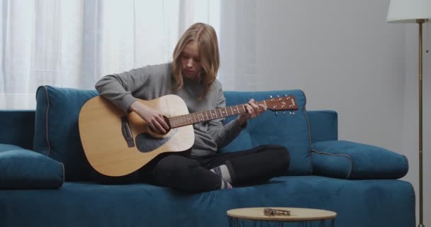 Una ragazza adolescente in abiti da casa suona una nuova melodia melodica su una chitarra classica si siede in una posizione comoda su un divano blu in un caldo soggiorno — Video Stock