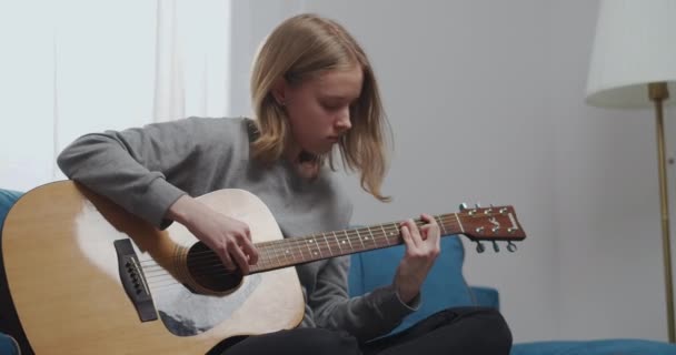 Een tiener in een grijs sweatshirt speelt gitaar terwijl hij op een blauwe bank zit. Middelmatig schot. — Stockvideo