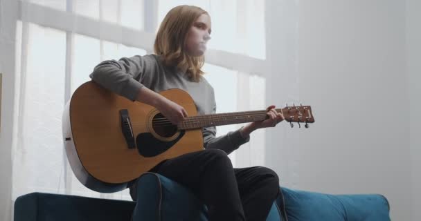 Девушка играет на гитаре на спинке дивана. Женщина создает музыку в помещении. Художник играет на акустической гитаре. Музыкант сочиняет мелодию. Подросток тренируется на музыкальном инструменте. — стоковое видео