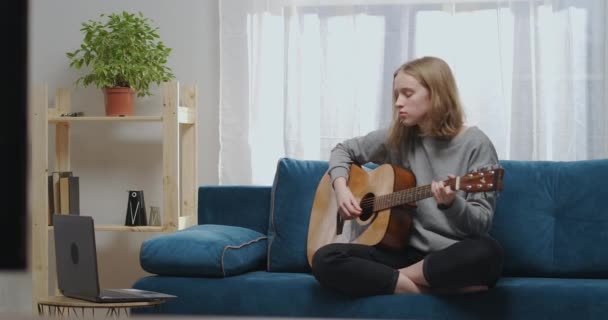 Nastolatek siedzi na miękkiej sofie. Młody muzyk uczy się grać na gitarze. Dziewczyna patrzy jak grać na instrumencie muzycznym na laptopie. — Wideo stockowe