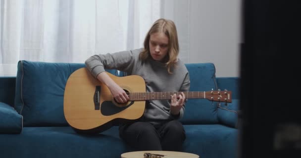 Egy zenész nyugodt dallamot játszik egy akusztikus gitáron, miközben egy lágy kék kanapén ül.. — Stock videók