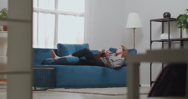 Attraktives junges Mädchen mit blonden Haaren ruht auf einem weichen Sofa in einem hellen Wohnzimmer. Der Musiker spielt klassische Gitarre. Weitschuss. — Stockvideo