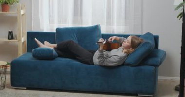 Oturma odasında gitar çalan çekici bir kadın hafta sonu yumuşak kanepede dinleniyor. Kız bacağını böyle müzikal bir melodiyle sallıyor..