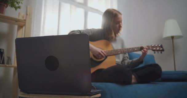 Joven chica hermosa se sienta en un sofá azul suave en una posición de loto en la sala de estar. Un adolescente aprende a tocar la guitarra con un portátil solo. Concéntrate en chica portátil en bokeh. — Vídeos de Stock