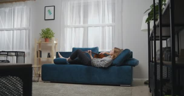 Vista lateral de uma jovem em roupas casuais deitada descalça em um sofá em uma sala de estar branca. Ela toca música romântica em uma guitarra acústica. Tiro longo — Vídeo de Stock